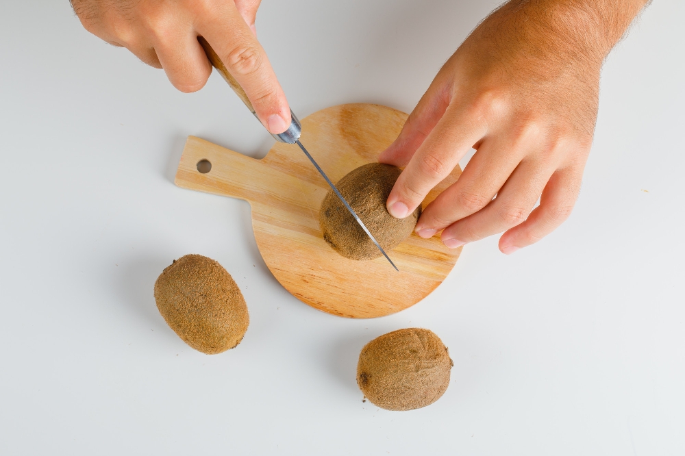 Flat lay of hands cutting a kiwi fruit on a wooden cutting board.